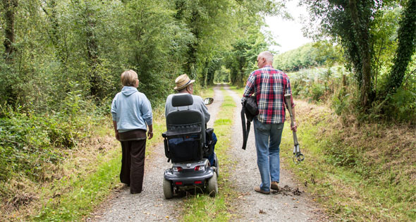 Three people walking down a leafy country lane, one on a mobility scooter